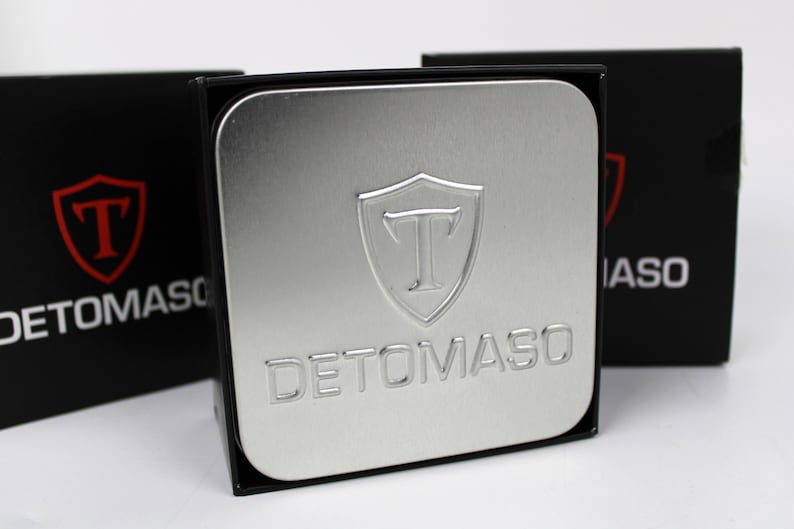 May include: A square silver metal tin featuring an embossed shield logo with the letter T and the text DETOMASO. The tin is positioned in front of two black boxes, each displaying a red shield logo with a T and the white text DETOMASO.