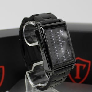 May include: A black rectangular wristwatch with a metal link band. The watch face features a vertical digital display with numbers 1 through 12 on the left and 05 through 55 on the right, with the text DEMASO on the side and a shield logo.