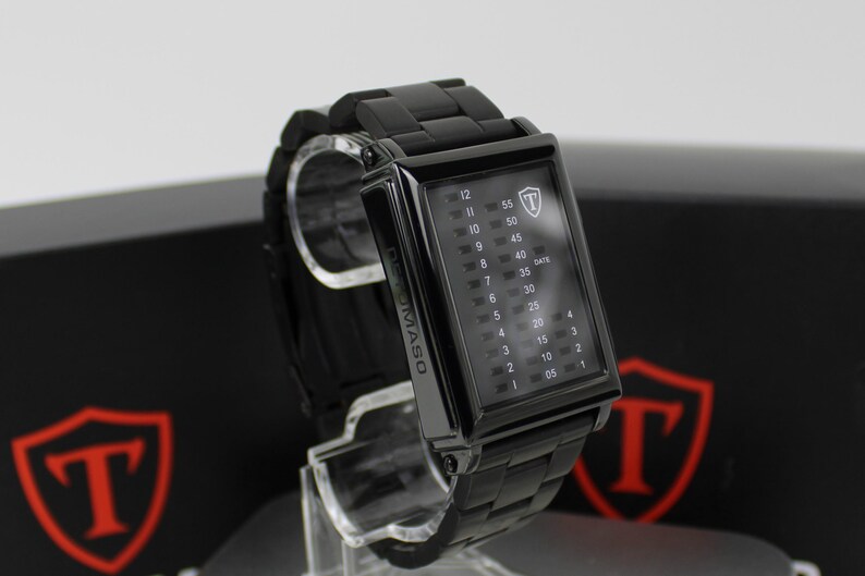 May include: A black rectangular wristwatch with a metal link band. The watch face features a vertical digital display with numbers 1 through 12 on the left and 05 through 55 on the right, with the text DEMASO on the side and a shield logo.