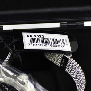 K&ouml;nnte beinhalten: Eine schwarze Uhrenbox mit einer silbernen Uhr darin. Die Uhr hat ein silbernes Metallband und ein schwarzes Zifferblatt. Ein schwarzes Etikett mit dem Text "XA.9522" und einem Barcode ist an der Box befestigt.