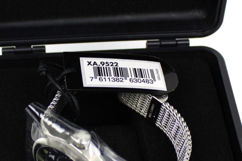 K&ouml;nnte beinhalten: Eine schwarze Uhrenbox mit einer silbernen Uhr darin. Die Uhr hat ein silbernes Metallband und ein schwarzes Zifferblatt. Ein schwarzes Etikett mit dem Text "XA.9522" und einem Barcode ist an der Box befestigt.