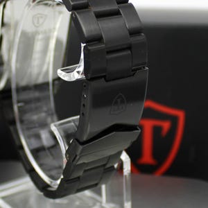 May include: A matte black metal watch with a link bracelet and a fold-over clasp. The clasp features an engraved shield logo with the letter T inside. The watch case is rectangular with a matching black finish and a clear protective film on the back.