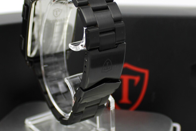 May include: A matte black metal watch with a link bracelet and a fold-over clasp. The clasp features an engraved shield logo with the letter T inside. The watch case is rectangular with a matching black finish and a clear protective film on the back.