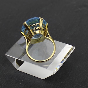 Puede incluir: Un anillo de oro con una gran piedra preciosa azul facetada. El anillo est&aacute; colocado en un expositor de acr&iacute;lico transparente.