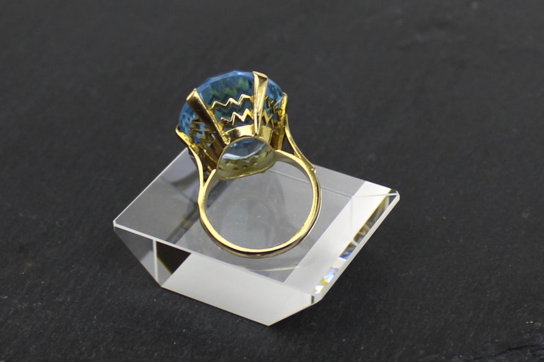 Puede incluir: Un anillo de oro con una gran piedra preciosa azul facetada. El anillo est&aacute; colocado en un expositor de acr&iacute;lico transparente.