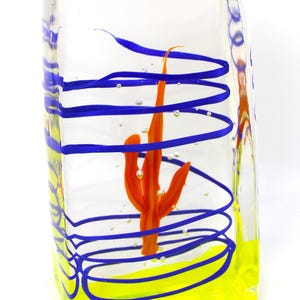 May include: A clear glass art piece with a bright orange cactus design inside. The glass is decorated with blue spiral lines and a yellow base. The piece is rectangular and has a unique, artistic appearance.
