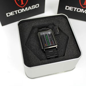 May include: A black Detomaso wristwatch with a rectangular face featuring a vertical digital display with green, yellow, and red light indicators. The watch has a black metal link band and is presented inside a square silver metal tin with foam.
