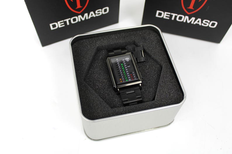 May include: A black Detomaso wristwatch with a rectangular face featuring a vertical digital display with green, yellow, and red light indicators. The watch has a black metal link band and is presented inside a square silver metal tin with foam.