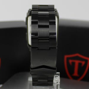 May include: A black metal wristwatch featuring a link bracelet with a deployment clasp. The watch band has a matte finish and is positioned vertically, showing the back of the clasp which displays a small embossed logo on the metal surface.