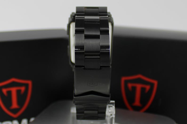 May include: A black metal wristwatch featuring a link bracelet with a deployment clasp. The watch band has a matte finish and is positioned vertically, showing the back of the clasp which displays a small embossed logo on the metal surface.