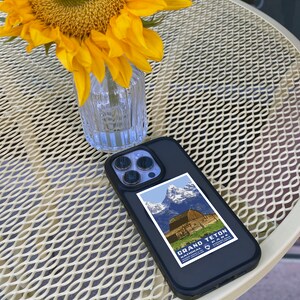May include: A black phone case with a vintage Grand Teton National Park poster design. The phone case is on a white metal table with a sunflower in a glass vase.