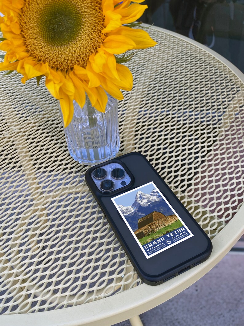 May include: A black phone case with a vintage Grand Teton National Park poster design. The phone case is on a white metal table with a sunflower in a glass vase.