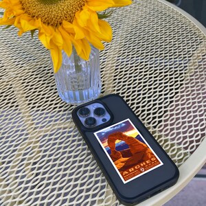 May include: A black iPhone case with a sticker of Arches National Park in Utah. The sticker features a sunset over Delicate Arch with the park's name and logo. The phone case is on a white metal table with a sunflower in a glass vase.
