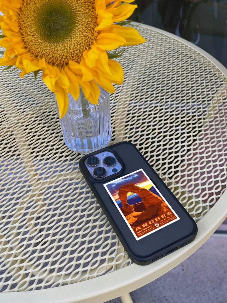 May include: A black iPhone case with a sticker of Arches National Park in Utah. The sticker features a sunset over Delicate Arch with the park's name and logo. The phone case is on a white metal table with a sunflower in a glass vase.