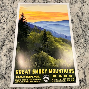 Op de afbeelding: Een vintage poster met een schilderachtig uitzicht op het Great Smoky Mountains National Park bij zonsondergang. De poster heeft een groene achtergrond met gele tekst die "Great Smoky Mountains National Park" en "Blue Ridge Mountains Appalachian Trail" linksonder en "Most Visited US National Park" rechtsonder staat.
