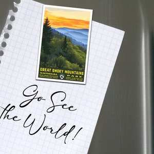 Op de afbeelding: Een wit vel papier met een rasterpatroon en een gescheurde rand. Op het papier staat de tekst "Go See the World!" geschreven in zwarte cursieve letters. Een vintage-achtige ansichtkaart met een zonsondergangsscene van het Great Smoky Mountains National Park is met een magneet aan het papier bevestigd.