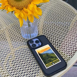 Op de afbeelding: Een zwarte iPhone-hoes met een sticker van een vintage reisposter voor het Great Smoky Mountains National Park. De sticker toont een zonsondergang boven een bergketen met bomen op de voorgrond. De telefoonhoes ligt op een witte metalen tafel met een zonnebloem in een glazen vaas.