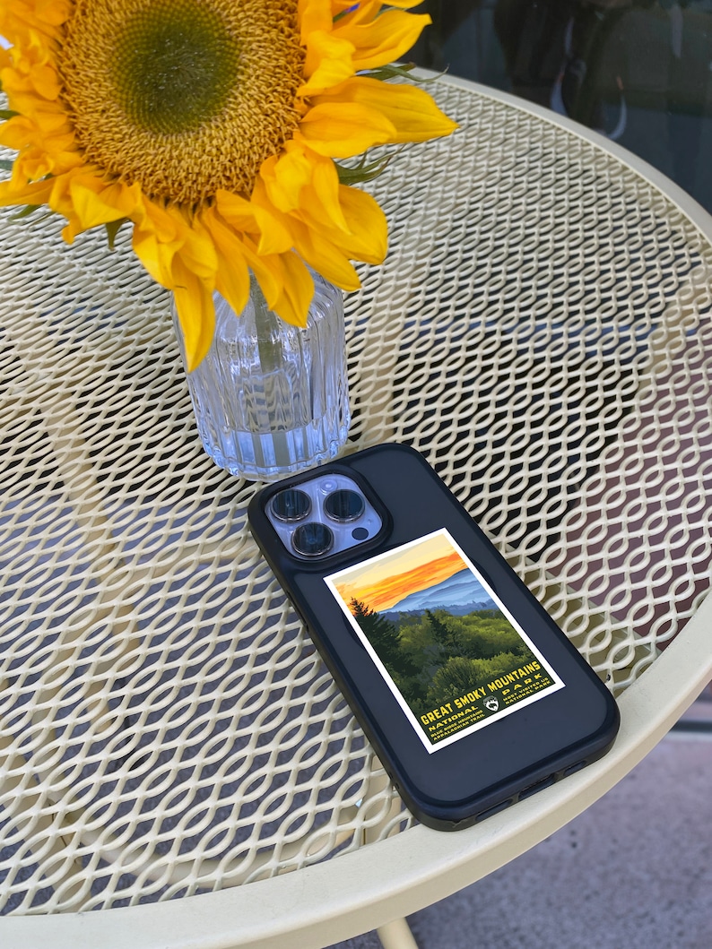 Op de afbeelding: Een zwarte iPhone-hoes met een sticker van een vintage reisposter voor het Great Smoky Mountains National Park. De sticker toont een zonsondergang boven een bergketen met bomen op de voorgrond. De telefoonhoes ligt op een witte metalen tafel met een zonnebloem in een glazen vaas.