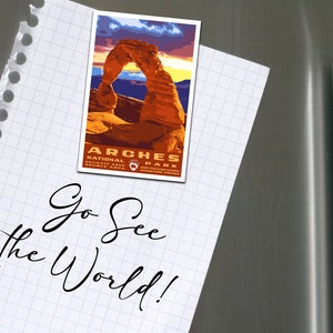 May include: A white notepad with a handwritten message that says "Go See the World!"  A magnet with a picture of Delicate Arch in Arches National Park is attached to the notepad.