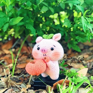 May include: A pink crocheted pig holding a peach. The pig has black eyes and a small green leaf on its head. The pig is sitting on a black base.