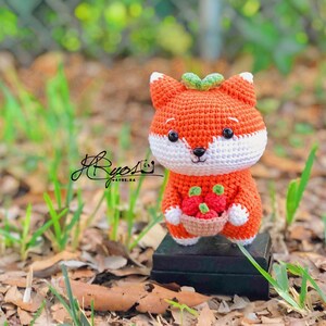 May include: A crocheted orange fox holding a basket of red strawberries. The fox has white fur on its face and chest. The fox is sitting on a black surface.