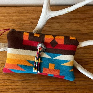 May include: A colorful, patterned zippered pouch with a southwestern design. The pouch features a brown top, a silver button with a beaded tassel, and a vibrant mix of orange, red, and blue geometric patterns. The pouch is resting on a white antler.