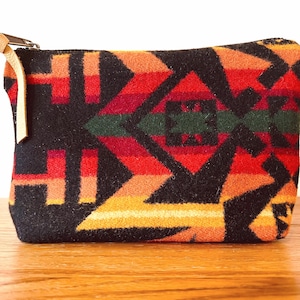 May include: A rectangular pouch with a colorful geometric pattern in shades of red, orange, yellow, green, and black. The pouch has a zipper closure with a tan leather pull tab. The pouch is made of a thick, woven fabric.