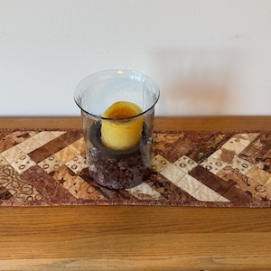 May include: A decorative table runner with a patchwork design in shades of brown and beige. A clear glass candle holder with a yellow candle sits on the runner. The runner is approximately 20 inches long.