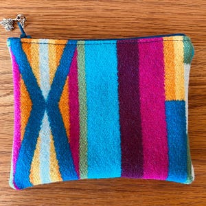 Peut inclure: Une pochette rectangulaire et colorée avec une fermeture éclair et un motif à rayures vives dans les tons de bleu, orange, rose et violet. Le motif comprend un motif bleu en forme de X. Un charm argenté est suspendu à la tirette.