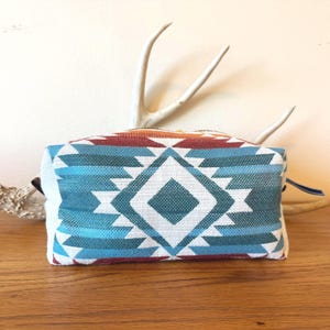 May include: A rectangular zippered pouch featuring a woven fabric exterior with a geometric pattern in shades of teal, light blue, white, and dark red. The design consists of a central diamond shape surrounded by stepped, angular motifs.