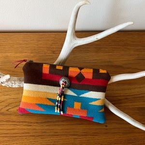 May include: A colorful, patterned zippered pouch with a southwestern design. The pouch features a brown top, a silver button with a beaded tassel, and a vibrant mix of orange, red, and blue geometric patterns. The pouch is resting on a white antler.