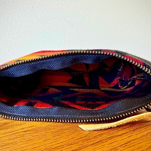 May include: Open zippered pouch with a colorful, patterned exterior. The pouch features a blue interior lining and a gold-colored zipper. A small tan leather pull tab is attached to the zipper.