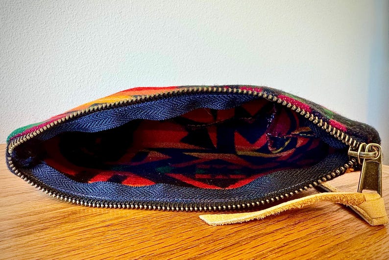 May include: Open zippered pouch with a colorful, patterned exterior. The pouch features a blue interior lining and a gold-colored zipper. A small tan leather pull tab is attached to the zipper.