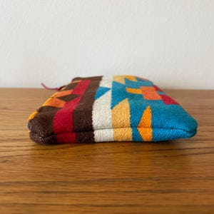 May include: A small, colorful pouch with a geometric pattern in shades of brown, red, orange, white, yellow, and blue. The pouch is made of a woven fabric and has a small loop at one end.