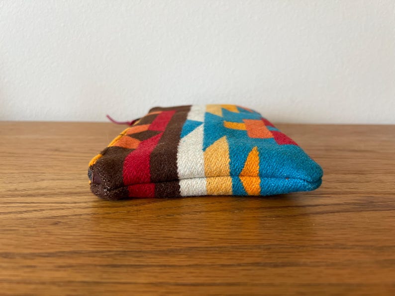 May include: A small, colorful pouch with a geometric pattern in shades of brown, red, orange, white, yellow, and blue. The pouch is made of a woven fabric and has a small loop at one end.