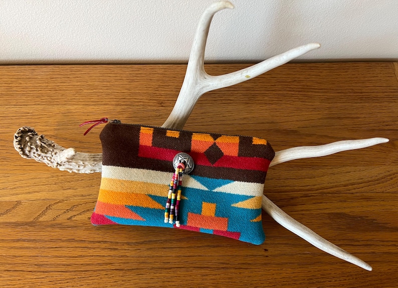 May include: A colorful, patterned zippered pouch with a southwestern design. The pouch features a brown top, a silver button with a beaded tassel, and a vibrant mix of orange, red, and blue geometric patterns. The pouch is resting on a white antler.