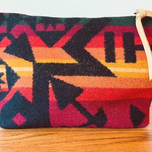 May include: A rectangular, patterned pouch with a leather loop. The pouch features a geometric design in shades of black, red, orange, yellow, and green. The leather loop is a light tan color. The pouch is likely made of a woven fabric.
