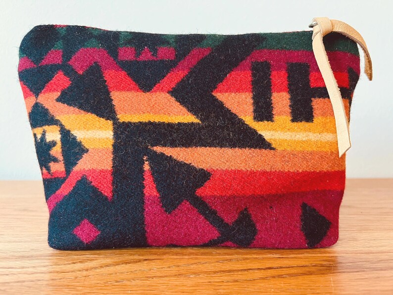 May include: A rectangular, patterned pouch with a leather loop. The pouch features a geometric design in shades of black, red, orange, yellow, and green. The leather loop is a light tan color. The pouch is likely made of a woven fabric.