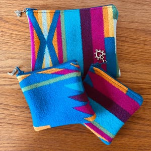 May include: Three colorful zippered pouches with a Southwestern-inspired design. The pouches feature a vibrant mix of blue, pink, orange, and yellow stripes and geometric patterns. Each pouch has a silver zipper pull and a decorative charm.