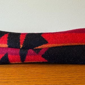 May include: A folded, rectangular textile with a geometric pattern in red, black, and orange. The fabric appears to be a woven material, possibly wool, with a repeating arrow-like design. The item is resting on a wooden surface.