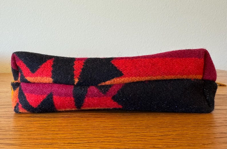 May include: A folded, rectangular textile with a geometric pattern in red, black, and orange. The fabric appears to be a woven material, possibly wool, with a repeating arrow-like design. The item is resting on a wooden surface.