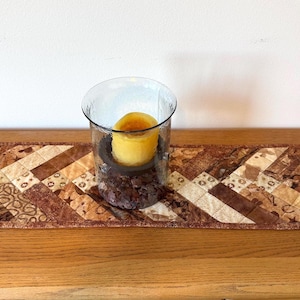 May include: A rectangular quilted table runner featuring a patchwork design in various shades of brown, tan, and cream. A clear glass cylindrical candle holder sits in the center, filled with dark brown decorative stones and a yellow pillar candle.