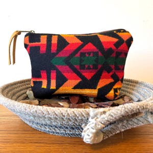 May include: A small, rectangular black pouch with a colorful geometric pattern in red, orange, green, and yellow. The pouch has a zipper closure and a tan leather wrist strap. It rests in a woven rope bowl.