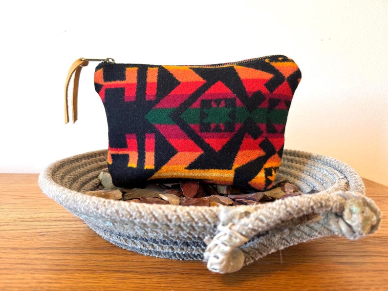 May include: A small, rectangular black pouch with a colorful geometric pattern in red, orange, green, and yellow. The pouch has a zipper closure and a tan leather wrist strap. It rests in a woven rope bowl.