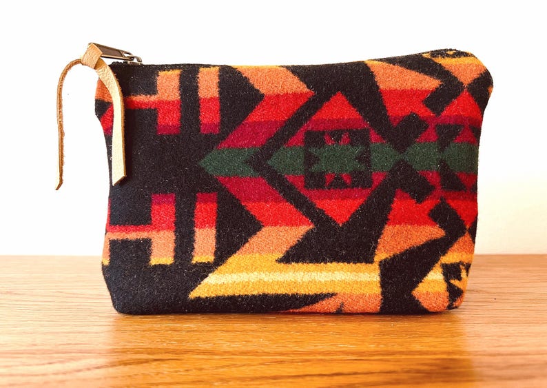 May include: A rectangular pouch with a colorful geometric pattern in shades of red, orange, yellow, green, and black. The pouch has a zipper closure with a tan leather pull tab. The pouch is made of a thick, woven fabric.