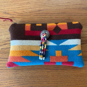 May include: A rectangular, patterned pouch with a zipper closure. The pouch features a brown top, a colorful geometric design in shades of blue, orange, red, and white, and a beaded tassel. A silver-toned button adorns the front.