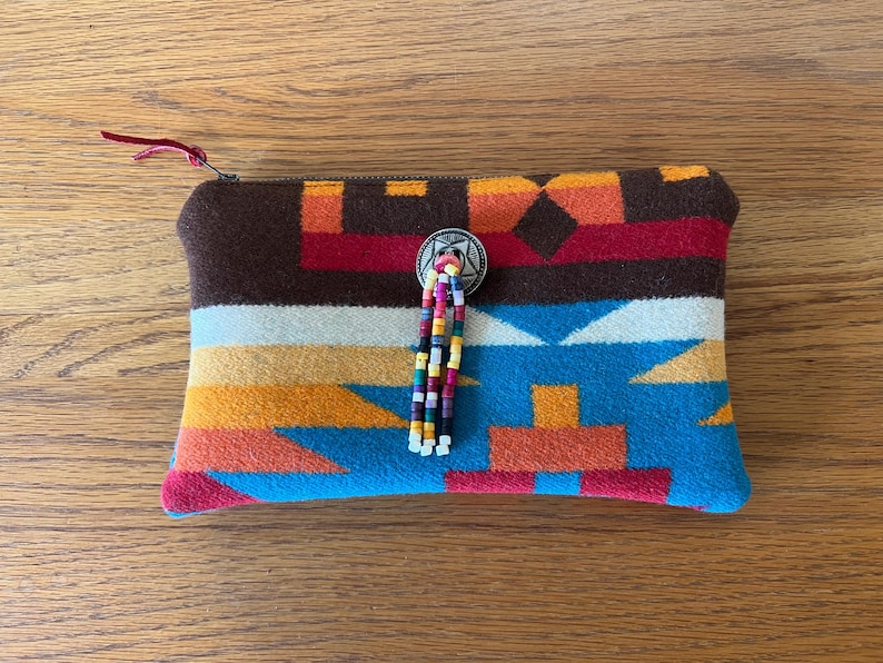 May include: A rectangular, patterned pouch with a zipper closure. The pouch features a brown top, a colorful geometric design in shades of blue, orange, red, and white, and a beaded tassel. A silver-toned button adorns the front.