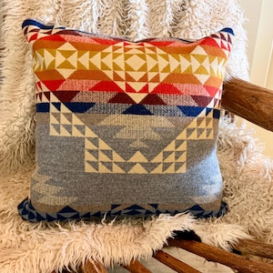 May include: A square throw pillow featuring a woven geometric pattern with a color gradient transitioning from deep red and orange at the top to a muted grey and blue at the bottom, showcasing intricate triangular and diamond-shaped motifs.