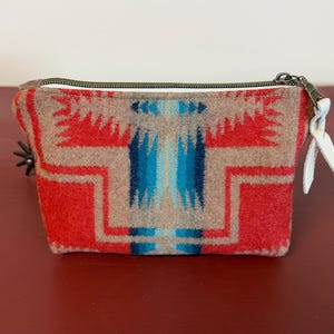 Può includere: Una pochette con cerniera caratterizzata da un motivo geometrico in rosso, beige e blu. La borsa presenta una chiusura a zip in metallo con una linguetta in pelle bianca e un piccolo ciondolo in metallo a forma di stella sul lato.