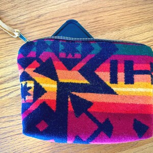 May include: A rectangular, zippered pouch with a colorful geometric pattern in shades of red, orange, yellow, green, and navy blue. The pouch has a leather wrist strap attached to the zipper.